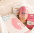 A curated gift box from Serein containing wellness products including a pink facial mask sheet, a pink can of Recess Candy, and a pink box of Earth Mama Nyonya Butter, arranged in white tissue paper inside a cream-colored box.